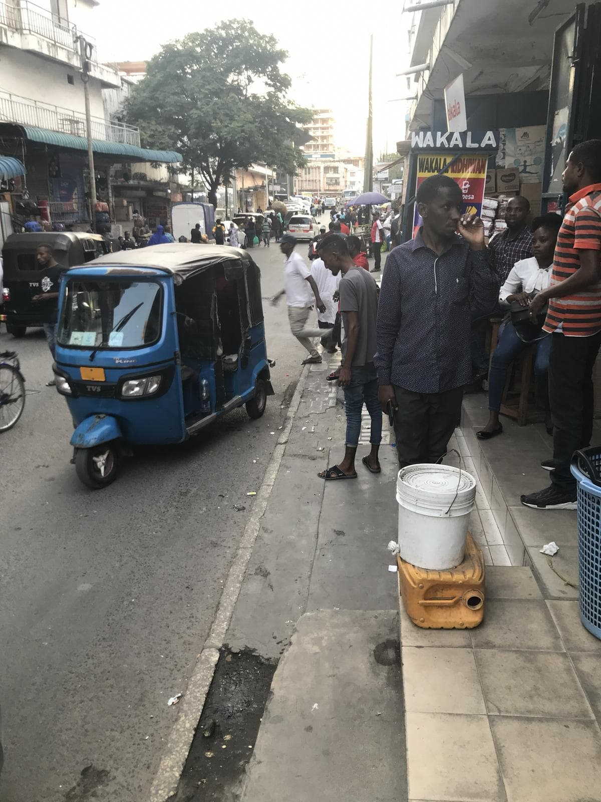 A blue bajaji parked on a Kariakoo street beside a mobile-money shopfront, people waiting on the pavement