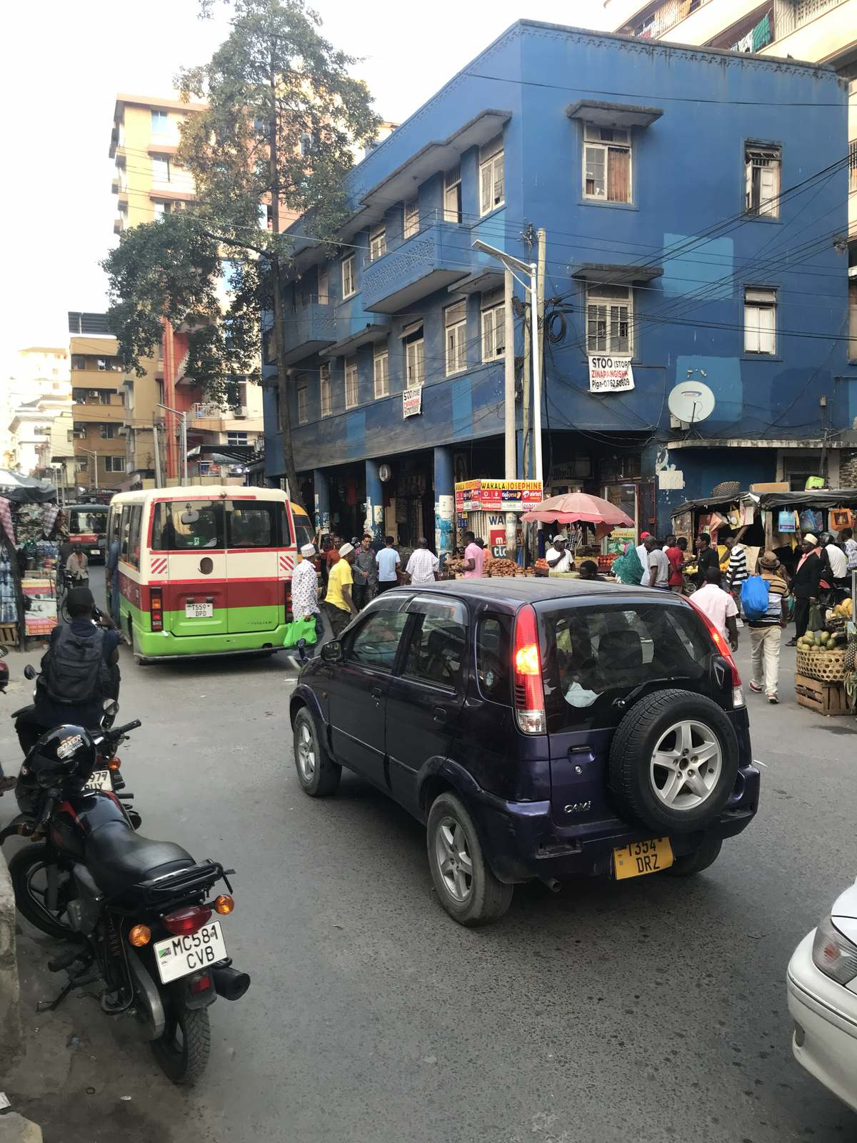 A daladala minibus passing a blue three-storey building on a Kariakoo corner, with fruit stalls and shoppers on the pavement