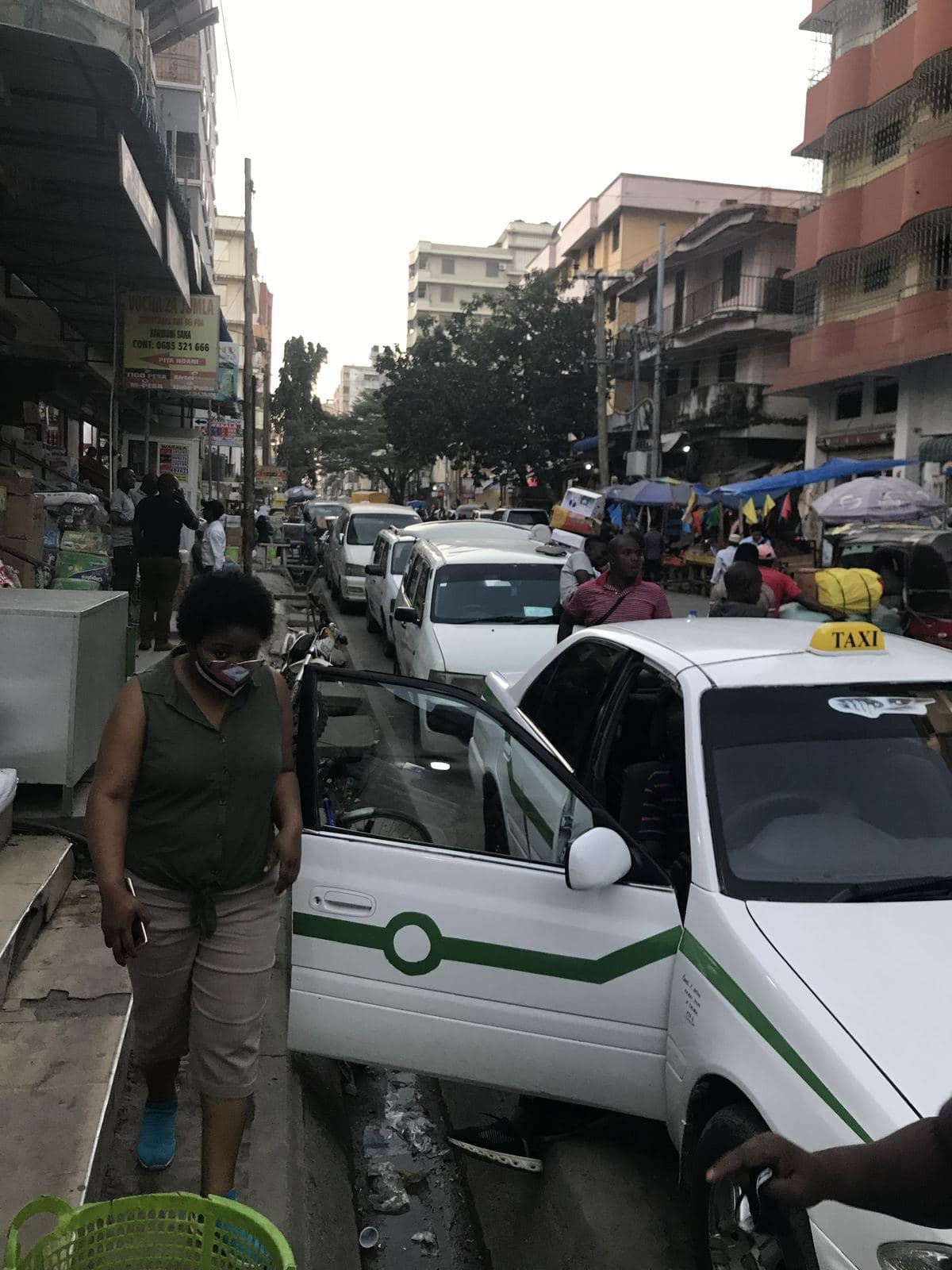 A taxi with its door open on a narrow Kariakoo street, parked cars ahead and market umbrellas along the pavement
